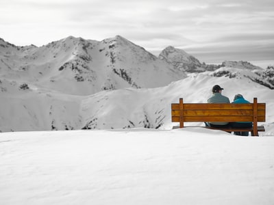 两个人坐在雪地上面对冰川山脉的长凳上